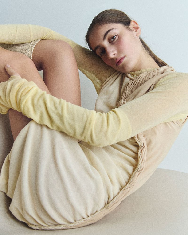 Young woman in pale yellow dress and top reclines indoors in a fashion shot with soft lighting and neutral studio environment.