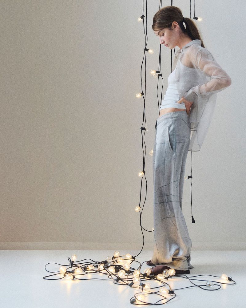 A stylish young woman stands near a string of lights indoors, wearing a semi-sheer top and grey trousers in a brightly lit setting.