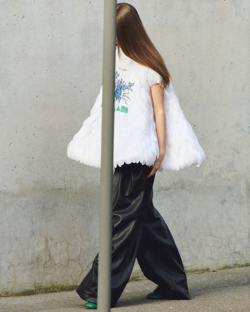 Fashionable person walking on a sidewalk wearing a white, floral top and dark pants with a concrete wall in the background.