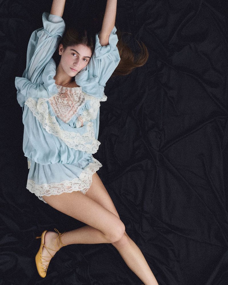 Overhead shot of a young woman in blue lace-trimmed loungewear and mustard-colored heels, posing on a black textile backdrop.