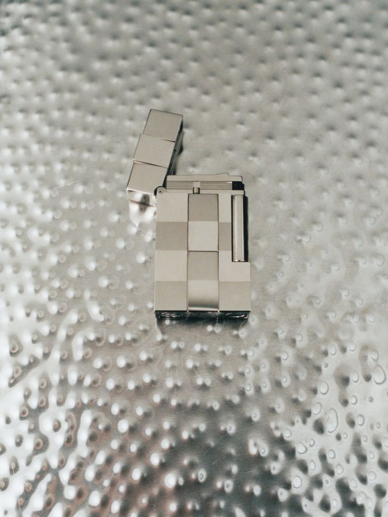 A silver metal lighter with a checkered pattern sits on a textured metallic surface in a high-angle shot with soft, diffused lighting.