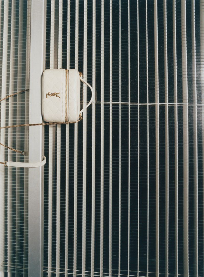 Ivory-colored Yves Saint Laurent purse with gold chain strap hanging against a textured metal wall with vertical bars and silver column.