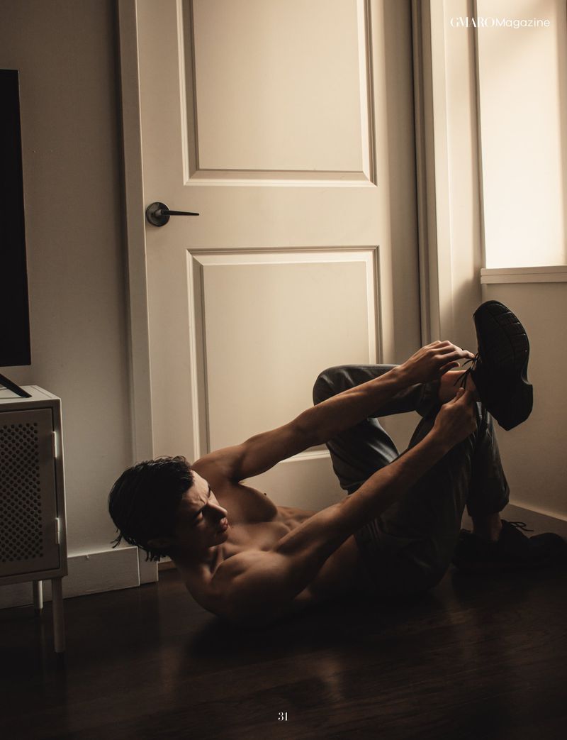 Muscular man in dark pants does abdominal crunches on wooden floor near a white door in a room with light walls and a small side table.