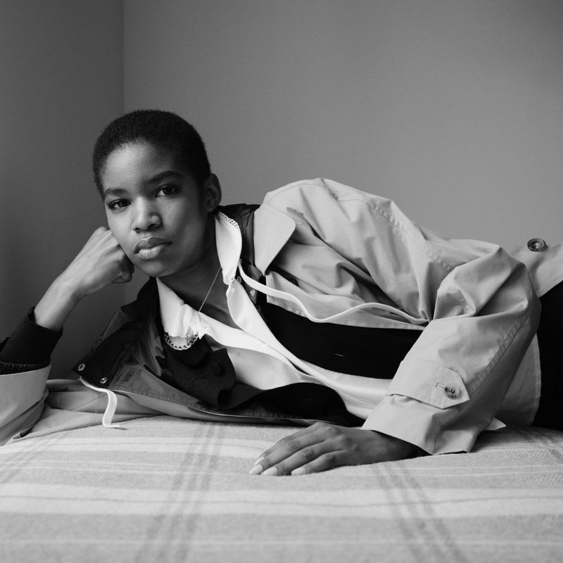 Black and white portrait of an African woman lying on a bed wearing a trench coat looking at the camera with her head propped on her hand.