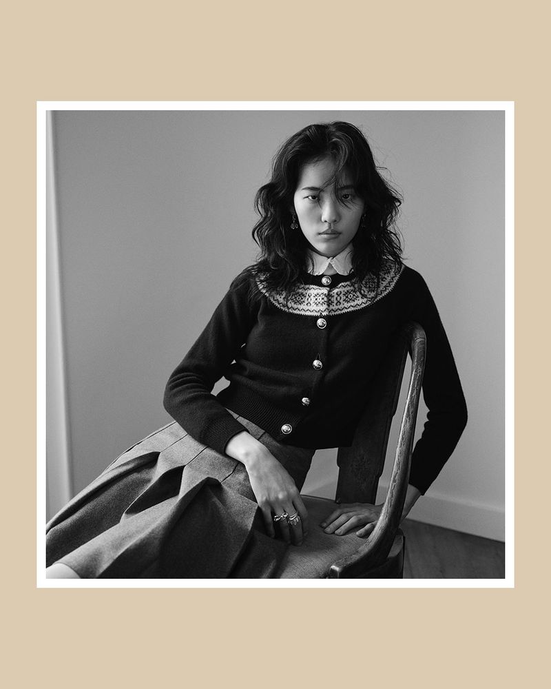 Black and white studio shot of a young Asian woman in a dark cardigan and pleated skirt, sitting on a chair against a plain wall.