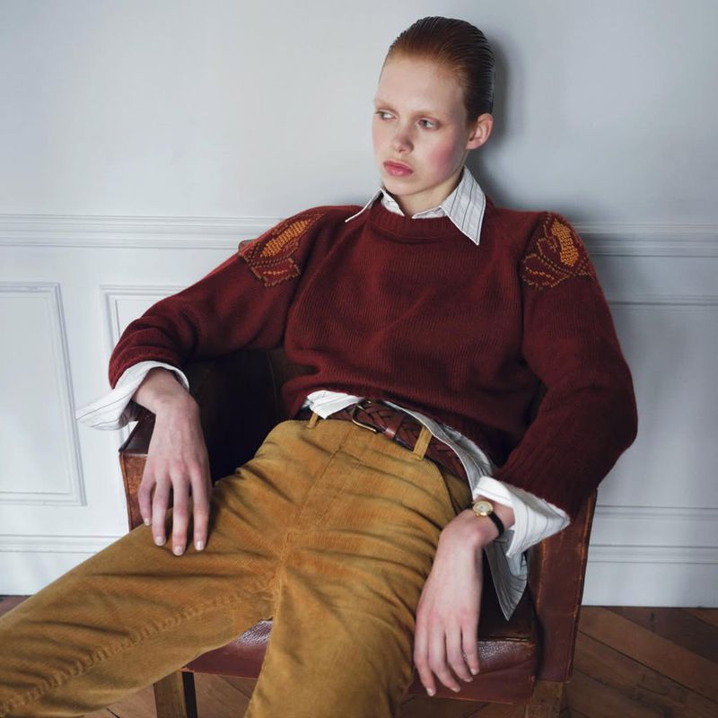 Model seated in an armchair wearing a red knitted sweater and yellow trousers against a white paneled wall