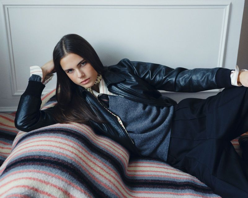Young woman with long brown hair poses while reclining on a striped blanket. She wears a black jacket, grey vest, and black pants indoors.