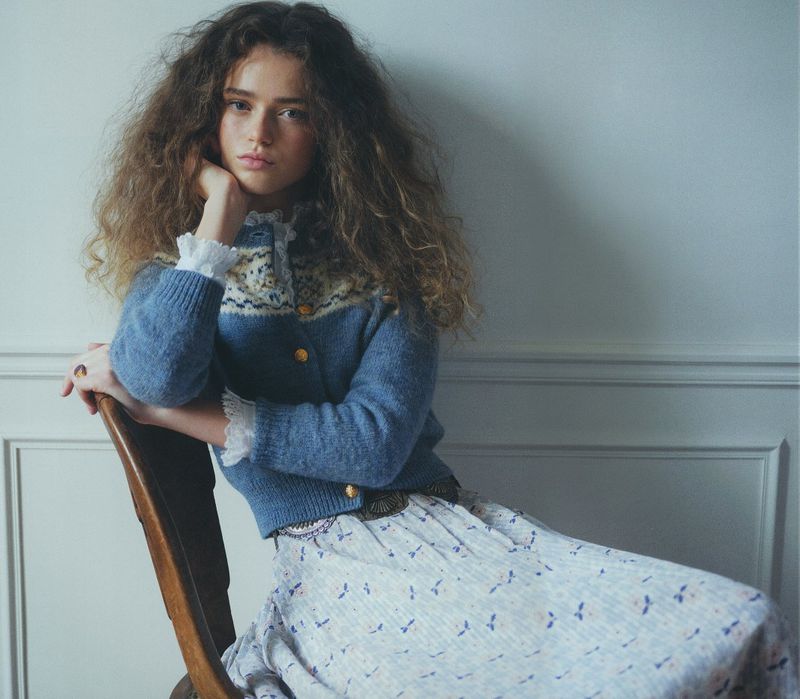 Young woman with curly hair sits on a chair, wearing a blue patterned cardigan and a floral print skirt in a soft, indoor setting.