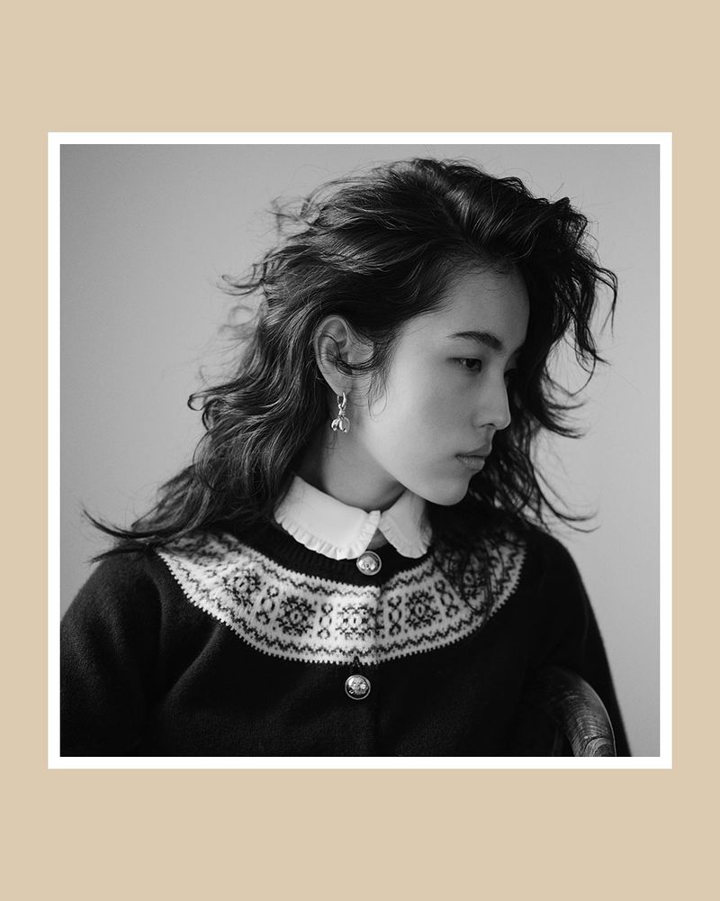 Black and white portrait of a young woman with wavy dark hair, wearing a patterned cardigan and earrings, against a neutral background.