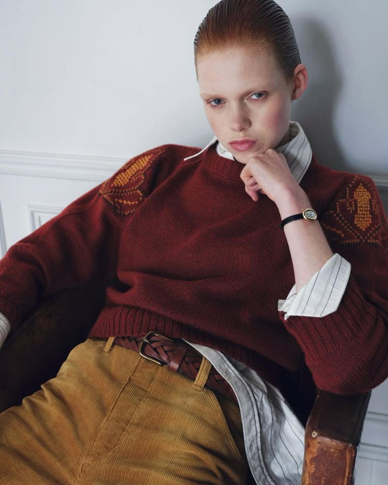 Portrait of a red-haired woman in a rust-colored sweater and corduroy pants, seated indoors with hand to face in a contemplative pose.