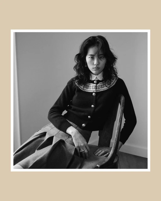 Model seated on a wooden chair wearing a dark patterned cardigan and pleated skirt in a black and white portrait.