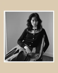 Model seated on a wooden chair wearing a dark patterned cardigan and pleated skirt in a black and white portrait.