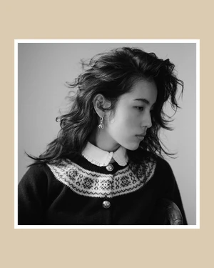 Monochrome portrait of a model with layered dark hair wearing a patterned button-up cardigan over a white collared shirt.