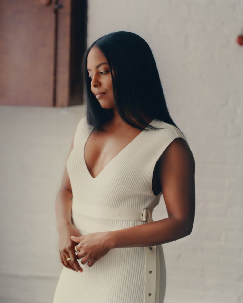 Black woman with long dark hair wearing a cream ribbed dress with a belt, standing indoors.