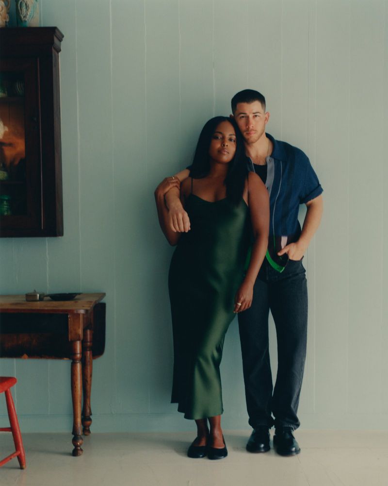 Two adults pose indoors against a light green wall, with a wood table, cabinet, and red stool providing background elements.