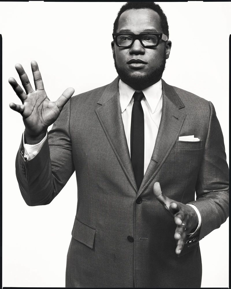 Black man in suit and glasses gestures against white background. Headshot features a tie, pocket square, and warm, monochrome tone.