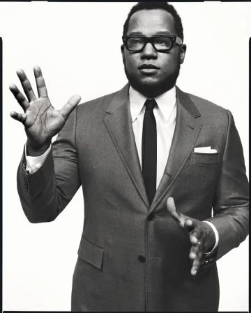 Black man in suit and glasses gestures against white background. Headshot features a tie, pocket square, and warm, monochrome tone.