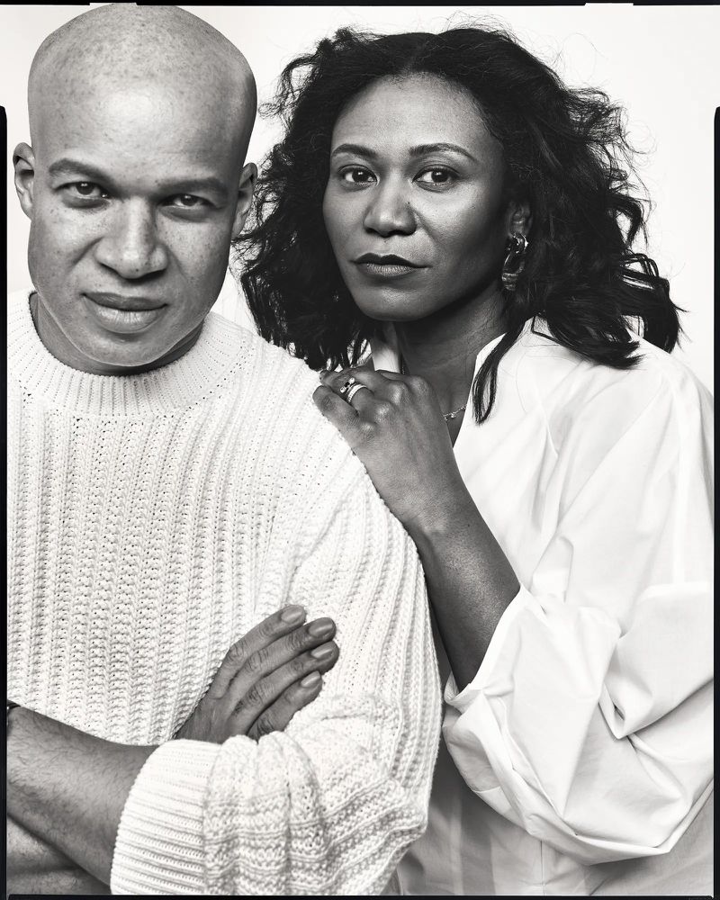 Monochrome portrait of an African American couple, the woman places a hand on the man's shoulder, posing indoors against a white backdrop.