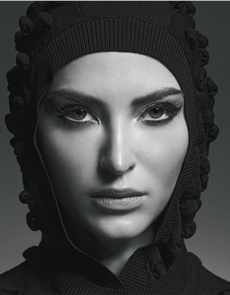 Close-up portrait of a woman with fair skin and dark eyes wearing a textured black hood. The lighting is soft, and the background is dark grey.