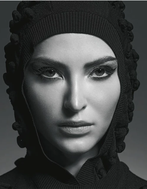 Close-up portrait of a woman with fair skin and dark eyes wearing a textured black hood. The lighting is soft, and the background is dark grey.