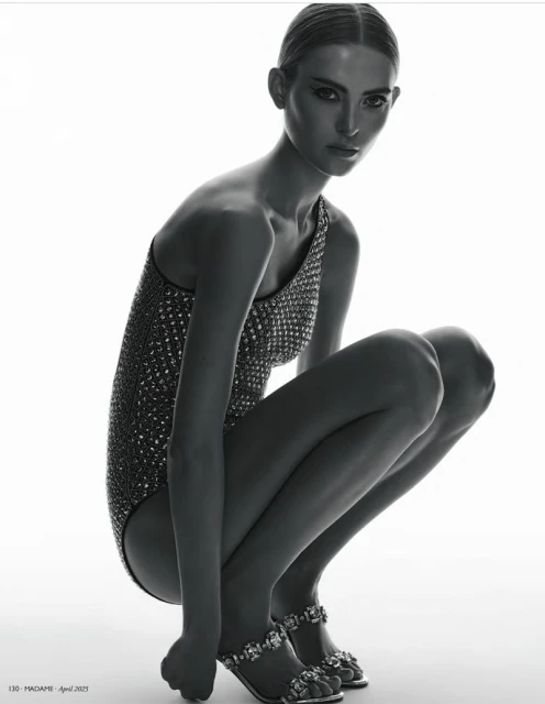 Monochrome fashion shot of a crouched woman in a glittering one-shoulder swimsuit and jeweled sandals against a bright white backdrop.