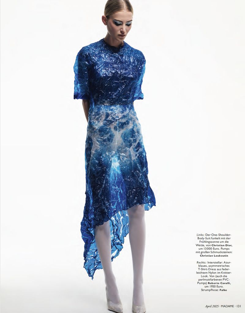 A young woman poses in a blue, translucent, high-fashion dress against a stark white backdrop in a studio setting.