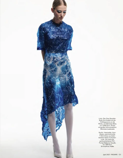 A young woman poses in a blue, translucent, high-fashion dress against a stark white backdrop in a studio setting.