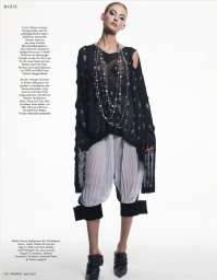 Model in black textured top and white pleated trousers with layered necklaces against a white background