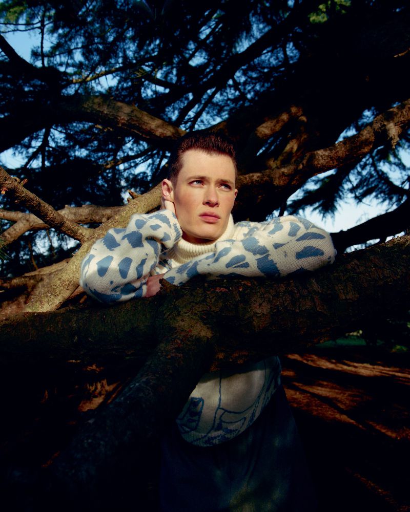 Portrait of a fair-skinned male model in a blue patterned sweater leaning against a pine tree branch in an outdoor setting.