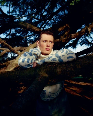 Portrait of a fair-skinned male model in a blue patterned sweater leaning against a pine tree branch in an outdoor setting.