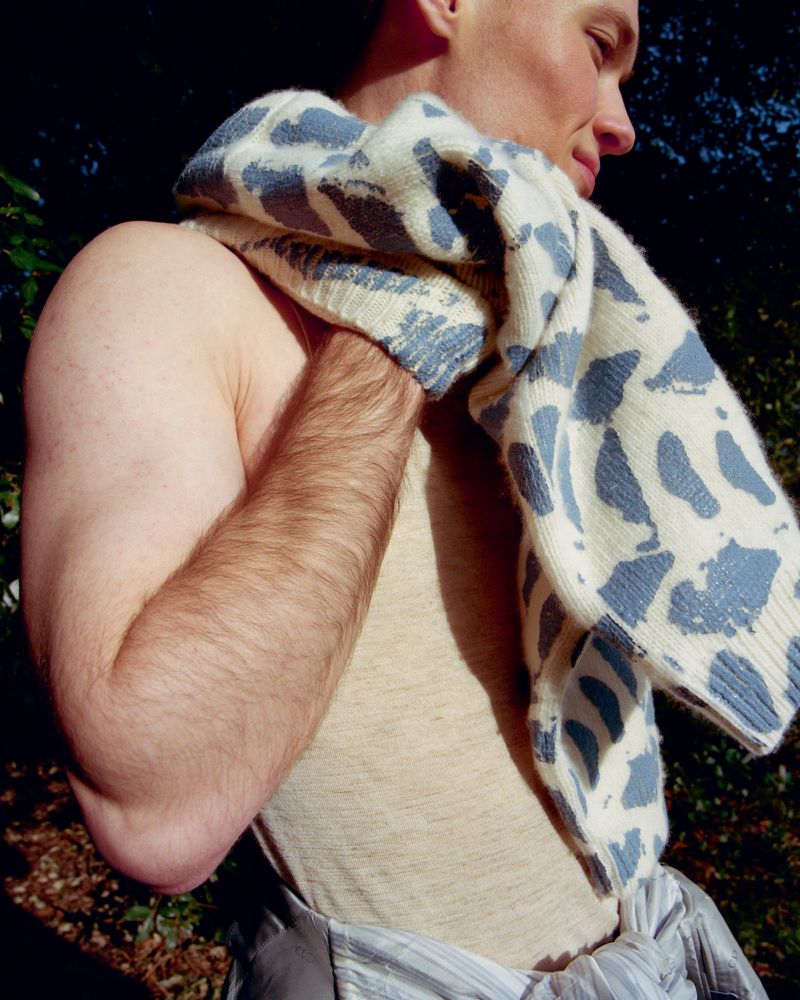 Adult male poses outdoors wearing patterned scarf around neck, neutral tank top, partial view of striped pants, trees in background.