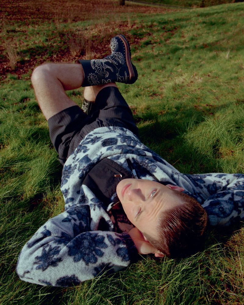 Person lying on green grass wearing floral patterned boots and jacket, casual shorts, bathed in warm sunlight, outdoor fashion.