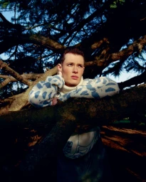 Model in a patterned blue and white turtleneck sweater leaning against a gnarled tree branch in an outdoor woodland setting.