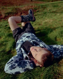 Model lying on grass wearing a patterned fleece pullover and embroidered boots for Fantastic Man Dior Special