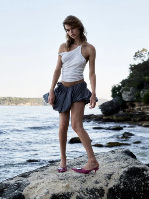 A slender woman with long hair poses on a rock by the ocean, wearing a white tank top, grey shorts, and pink heeled shoes in an outdoor setting.