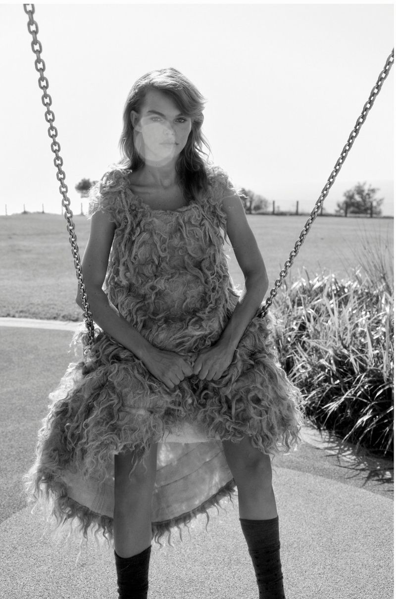 A woman in a textured dress sits on a swing in black and white, with a grassy field and trees in the background on a bright day.