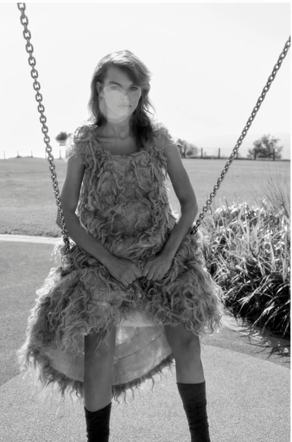 A woman in a textured dress sits on a swing in black and white, with a grassy field and trees in the background on a bright day.