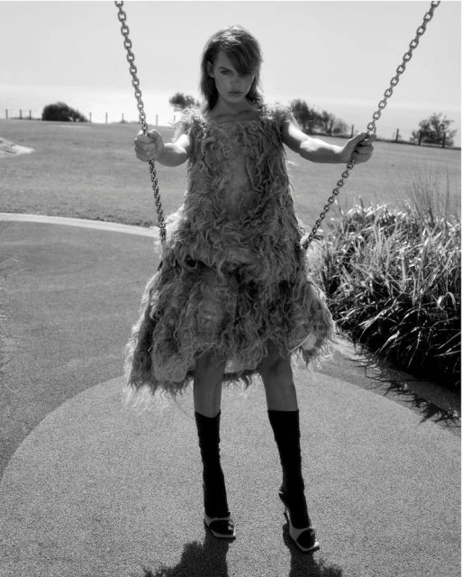 Black and white shot of a young woman in a feathered dress on a swing set in a park setting, looking towards the camera.