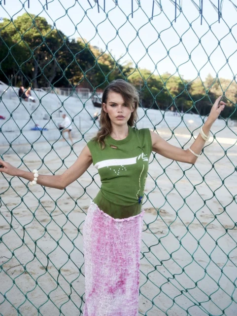 A woman in a green shirt and pink skirt stands behind a green chain-link fence at the beach on a sunny day, fashion shoot.