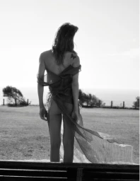 Monochrome editorial image of a model wearing a sheer draped garment outdoors against a horizon
