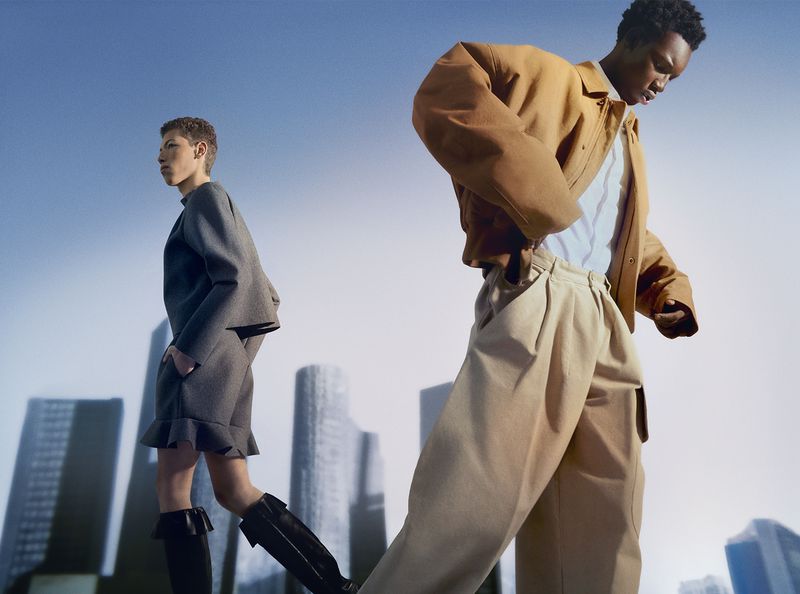 A low angle shot featuring two fashion models in a studio, showcasing a modern urban look with structured clothing and a backdrop of city buildings.