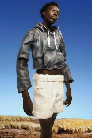 A young Black man wearing a grey textured hoodie and white shorts outdoors against a blue sky and barren landscape.