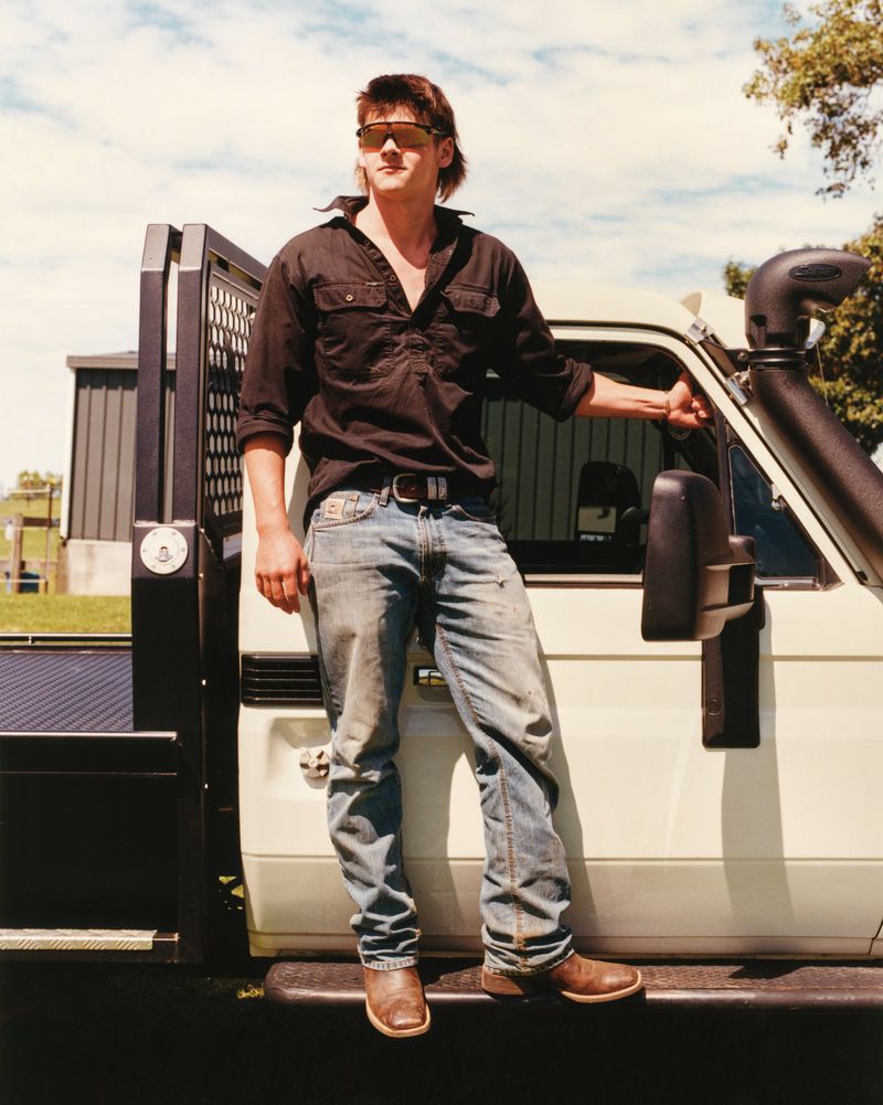 Adult male in sunglasses and dark shirt leans against a white pickup truck outdoors on a sunny day in a rural environment.