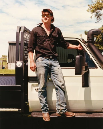 Adult male in sunglasses and dark shirt leans against a white pickup truck outdoors on a sunny day in a rural environment.