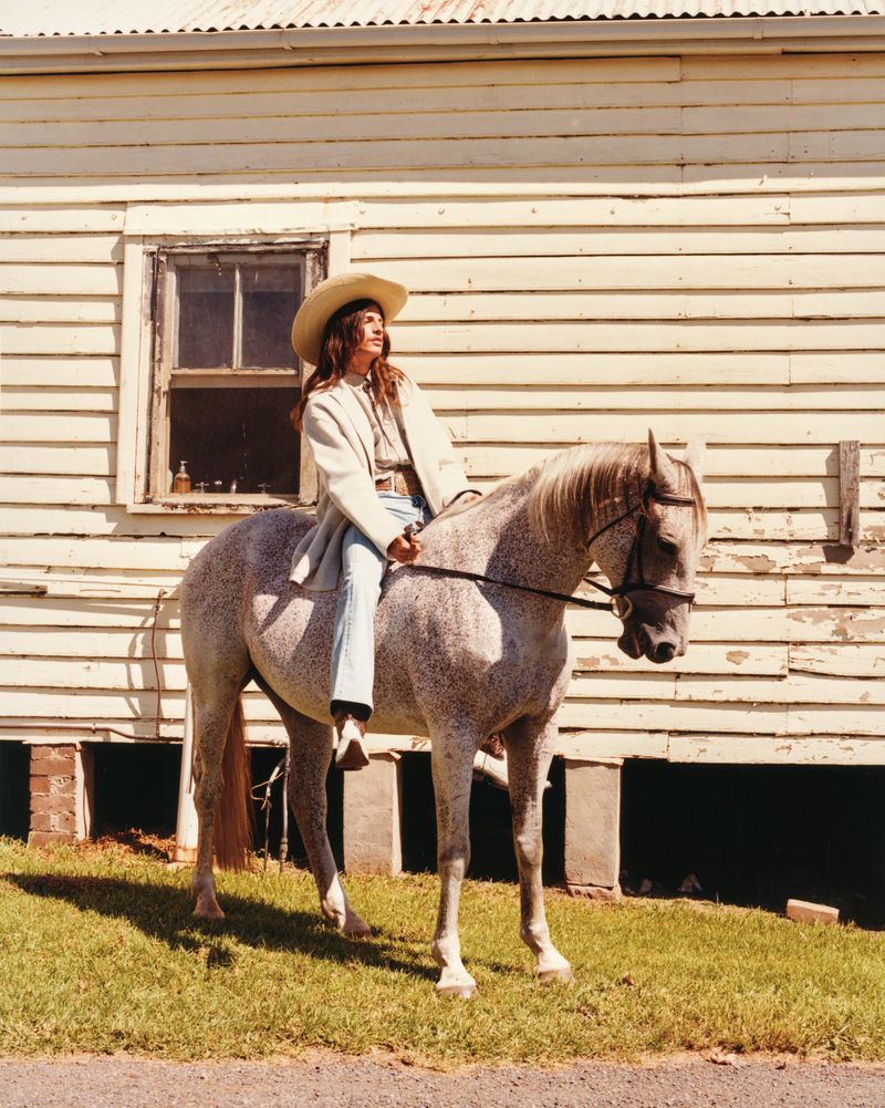 A woman in cowboy hat riding a dapple gray horse in front of a weathered building with a shingle roof and overgrown lawn.
