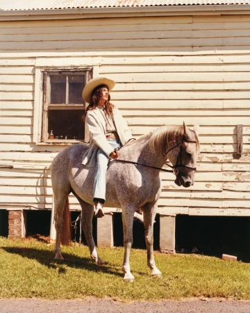 A woman in cowboy hat riding a dapple gray horse in front of a weathered building with a shingle roof and overgrown lawn.