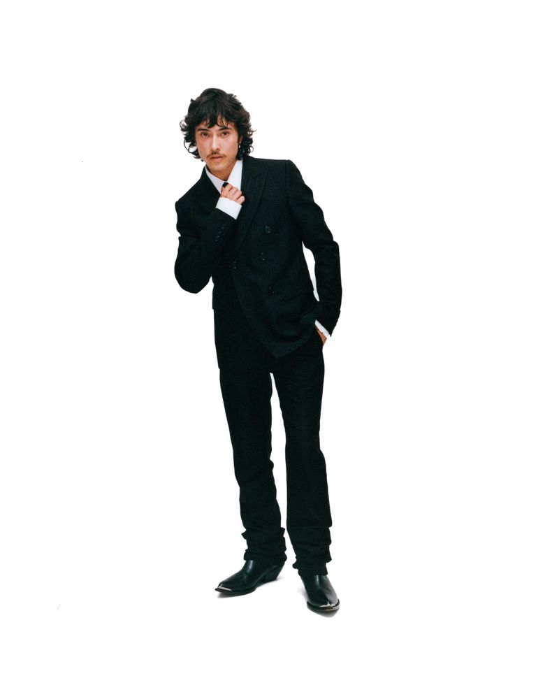 Full-length portrait of a young adult male in a black double-breasted suit with a white shirt, posing against a white background.
