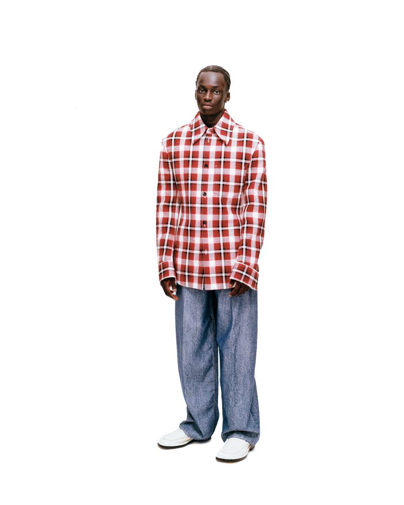 An adult male fashion model with dark skin and dark hair stands wearing a plaid shirt, denim pants, and white shoes on a white backdrop.