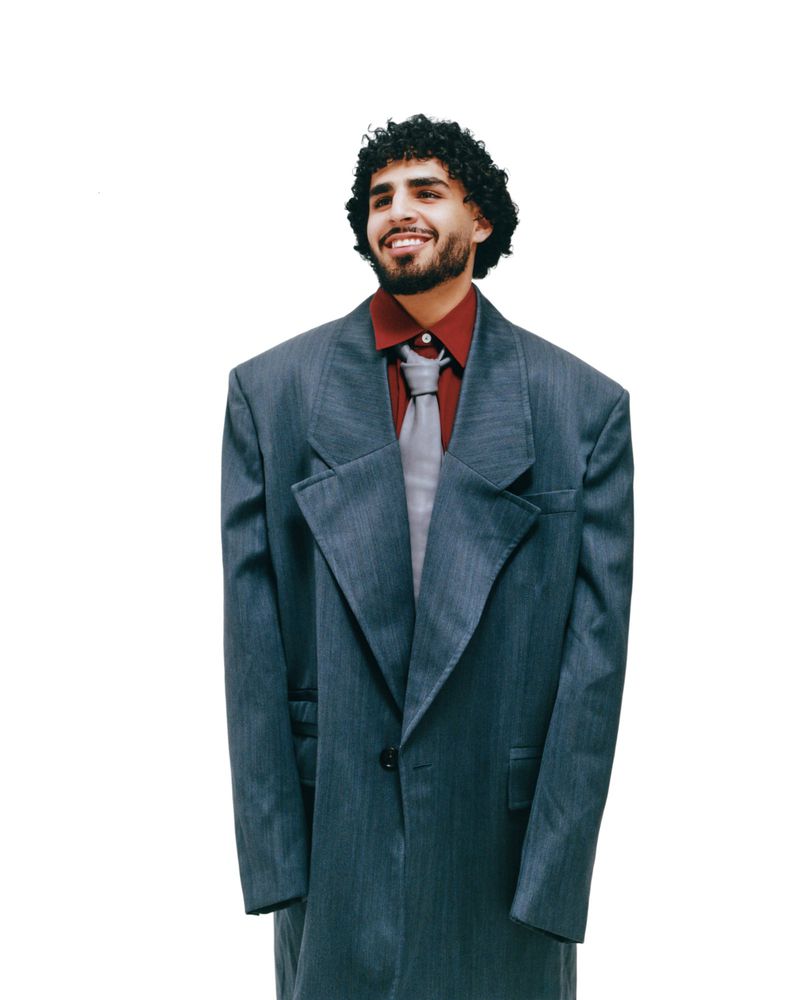 Portrait of smiling man with curly hair, wearing a red shirt, gray tie, and oversized blue-gray suit against a white backdrop.