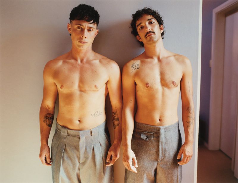 Two shirtless men pose indoors against a grey wall; both have tattoos and are wearing grey pants. One man has a mustache.